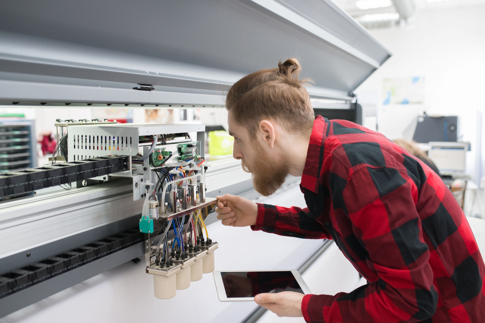 Serious concentrated hipster engineer with beard wearing casual shirt examining printing mechanism of machine and using tablet for notes in printing house